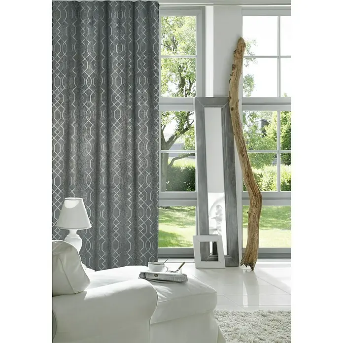 Elbersdrucke Verdunkelungsvorhang Equinox - 140 x 255 cm, 100 % Polyester, Anthrazit - Grauer Vorhang mit geometrischem Muster hängt im Wohnzimmer. Dazu gibt es ein weißes Sofa, eine Lampe und Fenster mit Blick ins Grüne.