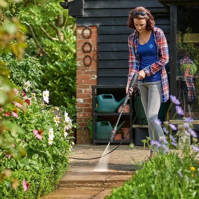 Bosch Hochdruckreiniger UniversalAquatak 130 - 1.700 W, Fördermenge: 380 l/h - Frau reinigt mit einem Hochdruckreiniger die Steinterrasse vor einem Gartenhaus. Das Gerät entfernt Schmutz mühelos.