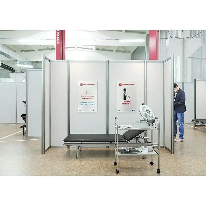 Hartschaumplatte ProtexWeiß, 50 cm x 50 cm x 3 mm, PVC - Impfzentrum mit Liege, Infusionswagen und Hinweisschildern für QR-Code und freiem Oberarm, während ein Mann mit Maske im Hintergrund steht.