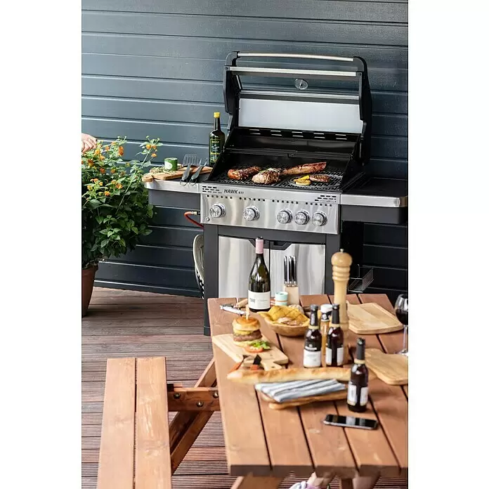 Grandstate Gasgrill Hawk 411 - Grillfläche (B x T): 71 x 49 cm, 15,2 kW, Anzahl Brenner: 4 Stk. - Ein Gasgrill aus Edelstahl mit schwarzem Gestell, der Fleisch und Gemüse auf einer Terrasse neben einem Esstisch zubereitet.