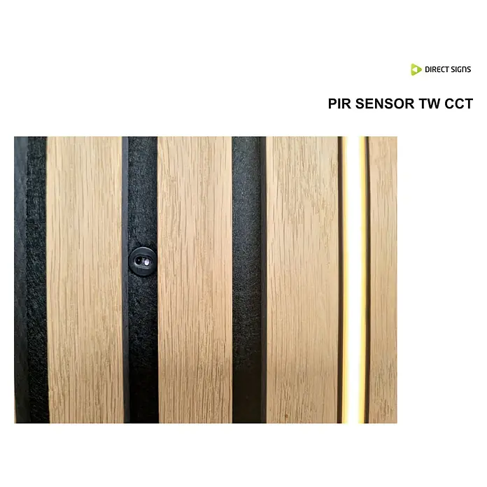 Direct Signs Sensor PIR CCT - Netzbetrieben, Schwarz, 2 x 1,2 x 2 cm - Nahaufnahme eines PIR-Sensors TW CCT, eingebettet in eine moderne Holzpaneelwand mit schlanken schwarzen Akzenten und Beleuchtung.