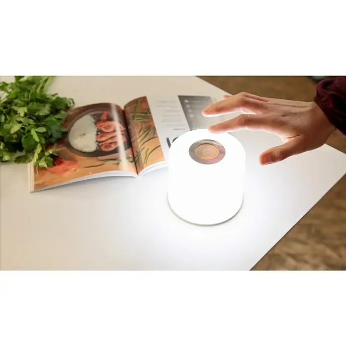 Lutec Akku-Tischleuchte NOMA Erweiterung2,3 W, RGBW, Farbe: Weiß, 1 Stk. - Eine Hand berührt eine moderne, runde Lampe auf einem Tisch, neben einem aufgeschlagenen Kochbuch und frischen Kräutern.