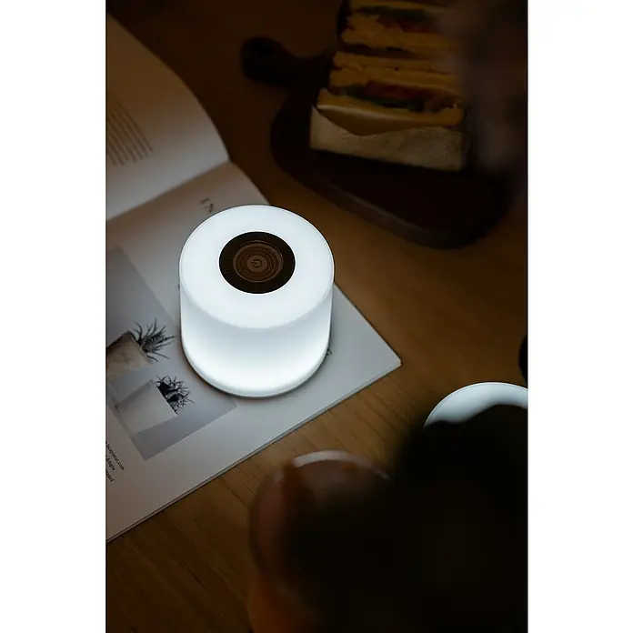 Lutec Akku-Tischleuchte NOMA Erweiterung2,3 W, RGBW, Farbe: Weiß, 1 Stk. - Nahaufnahme einer Tischlampe mit sanftem Licht, die auf einem offenen Buch steht, und einem Sandwich auf einem Holzbrett im Hintergrund.