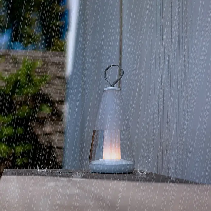 Lutec Akku-Tischleuchte PEPPER - 3 W, L x B x H: 12 x 12 x 29 cm, Weiß, RGBW - Diese Aufnahme zeigt eine wasserdichte Lampe im Regen, die eine warme und gemütliche Atmosphäre schafft.