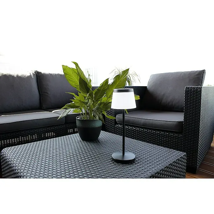 Lutec Akku-Tischleuchte OLEG - 1,5 W, Schwarz, Warmweiß - Nahaufnahme einer Tischlampe auf einem Terrassentisch, umgeben von einer Zimmerpflanze und einem modularen Lounge-Set aus Polyrattan.