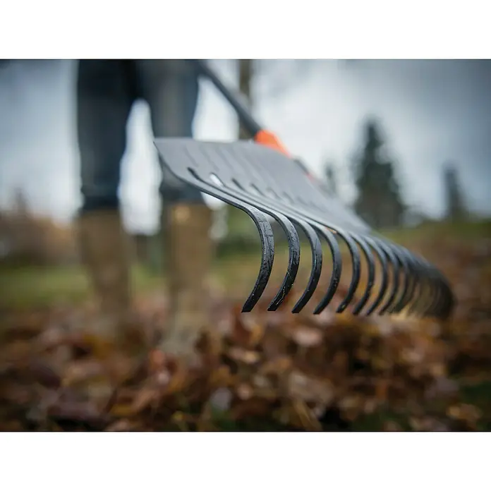Fiskars Solid Laubbesen - Arbeitsbreite: 52 cm, Anzahl Zinken: 24, Art Stiel: Langstiel - Nahaufnahme einer schwarzen Gartenharke, die über Herbstlaub gehalten wird, mit einer Person im Hintergrund.