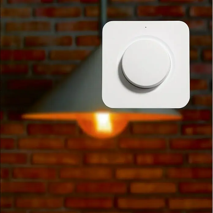 Intertechno Funk-Wandsender Drehdimmer-Schalter - Reichweite ca.: 30 m - Nahaufnahme eines weißen Smart Home-Dimmers vor einer Backsteinmauer und einer Lampe.