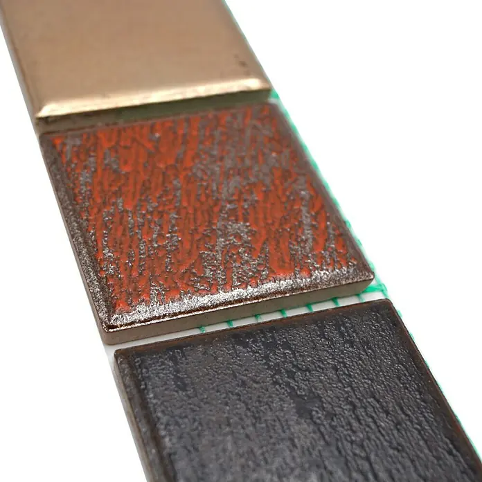 Fliesenbordüre Orient Color YBO CDAUBIS - 5 x 31,6 cm, Mix Braun/Grau, Matt - Nahaufnahme von drei quadratischen Keramikfliesen in Gold, Orange und Schwarz, die auf einem weißen Untergrund liegen.