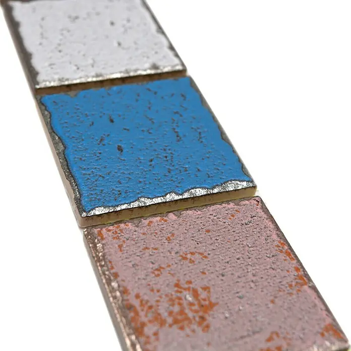Fliesenbordüre Orient Color YBO CDCUBIS - 5 x 31,6 cm, Bunt, Matt - Nahaufnahme von drei emaillierten Metallproben in Weiß, Blau und Rosa, die auf möglichen Oberflächenverschleiß hinweisen.