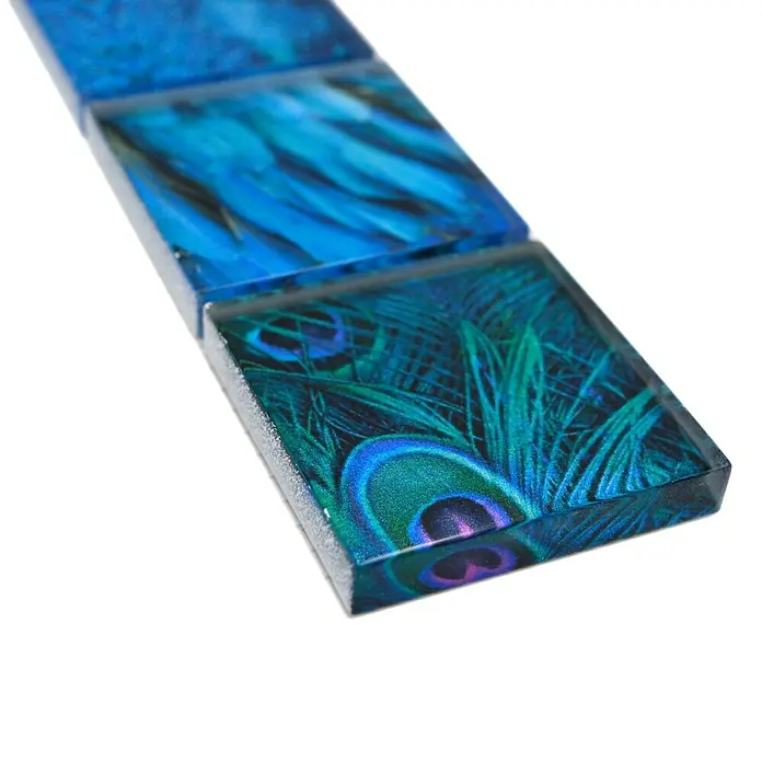 Fliesenbordüre Wildlife YBO WL78 - 29,8 x 4,8 cm, Blau, Glänzend - Nahaufnahme von drei blauen Glasmosaikfliesen mit Pfauenfederdesign, die auf einer reflektierenden Oberfläche liegen.