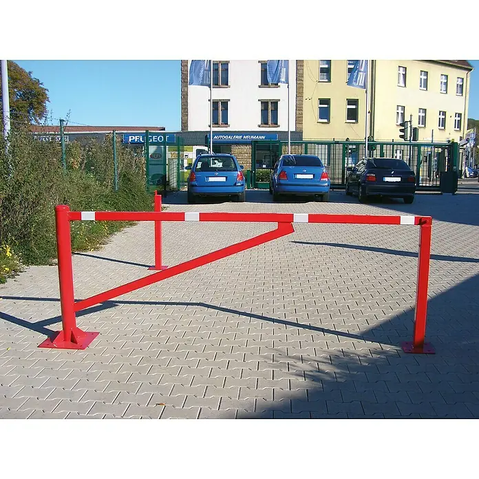 Schake Drehschranke - Mit Diagonalverstrebung, Art Schloss: Profilzylinderschloss, Länge: 4,5 m - Ein roter Schrankenbaum mit weißen Streifen versperrt die Einfahrt. Im Hintergrund sind geparkte Autos und ein Gebäude zu sehen.