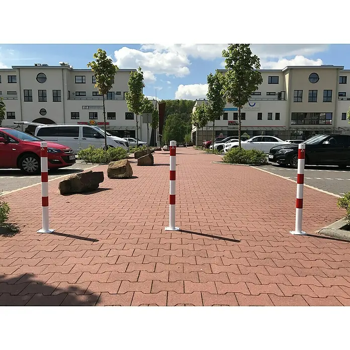 Schake Sperrpfosten - Ø x H: 60 x 900 mm, Stahl, Aufdübeln, Weiß/Rot, Öse - Rot-weiß gestreifte Sicherheitspoller und Felsbrocken auf einem Backsteinplatz, die Bereiche abgrenzen und vor Fahrzeugen schützen.
