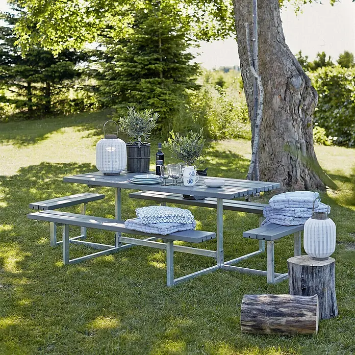 Plus Picknicktisch Basic - Graubraun, Kiefer/Fichte & Stahl, 8 Personen, ohne Rückenlehne - Eine graue Picknicktischgarnitur mit Holzbänken und Metallrahmen steht auf einer grünen Wiese, dekoriert mit Laternen, Pflanzen und Geschirr.