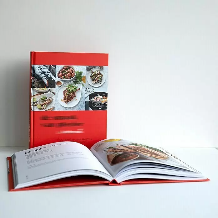 Barbecook Grillkochbuch: So schmeckt Spaß! -  - Zwei rote Kochbücher, eines steht geschlossen, das andere liegt offen mit Rezepten und Essensfotos.