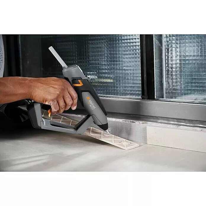 Steinel Heißklebepistole PRO GlueMatic 7011 - Durchmesser Klebepatrone: 11 mm, Temperaturbereich: 40 °C - 230 °C - Hand trägt Heißkleber mit grau-schwarzer Steinel PRO GM 7011 Klebepistole auf eine Fliese auf.
