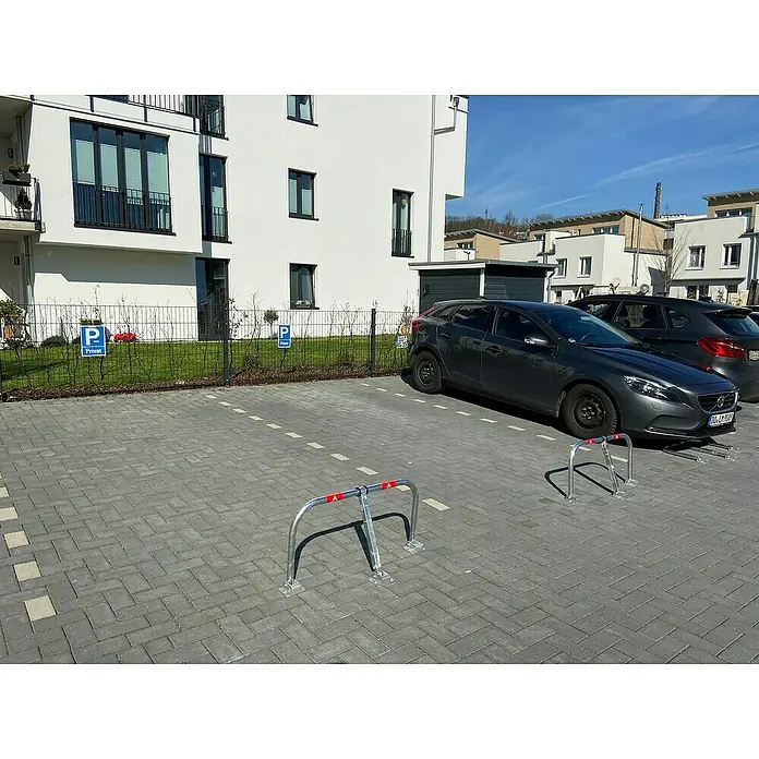 Schake Sperrbügel - L x B x H: 850 x 33,7 x 450 mm, Stahl, Aufdübeln, Profilzylinderschloss - Eine graue Volvo V40 parkt auf einem Parkplatz vor einem modernen Wohngebäude mit Fahrradständer.