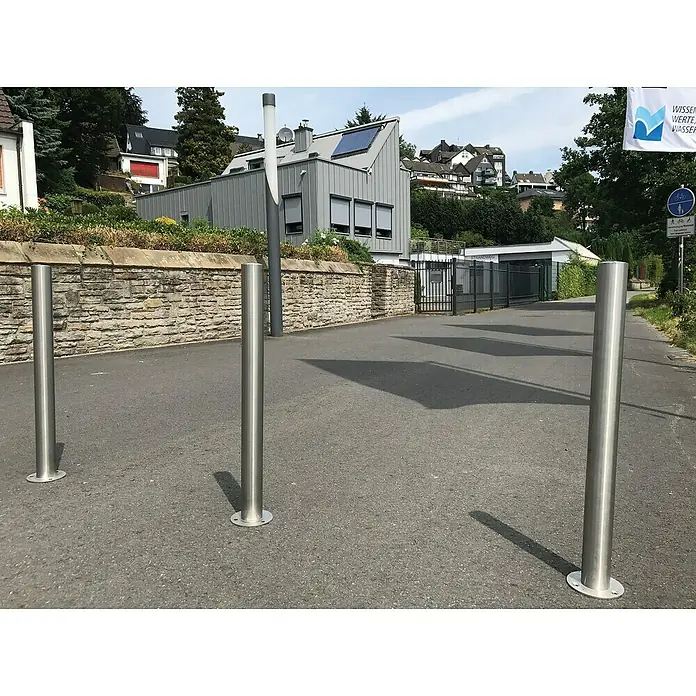 Schake Sperrpfosten - Ø x H: 60 x 1.300 mm, Edelstahl, Einbetonieren, Silber, Profilzylinderschloss - Straßenbild mit drei Edelstahlpfosten, die den Weg begrenzen und Sicherheit bieten, vor dem Hintergrund einer Stadtlandschaft.