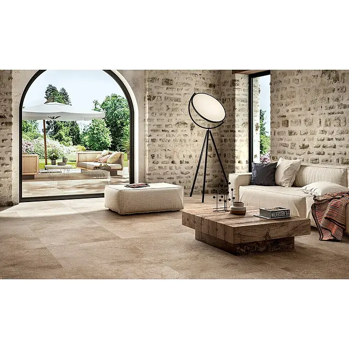 Marazzi Feinsteinzeugfliese Jerusalem - 75 x 75 cm, Noce, Matt - Einladendes Wohnzimmer mit Fliesenboden in Naturfarben, das nahtlos in eine grüne Gartenoase übergeht. Moderne Lampe und rustikaler Tisch ergänzen das Interieur.
