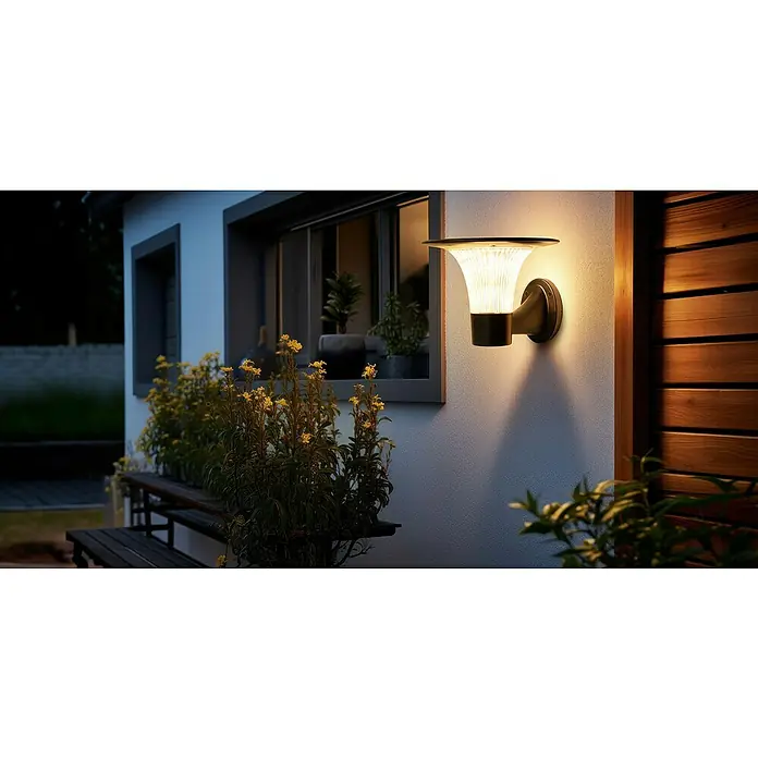 Garza Goliat Aplique solar para exterior con sensor - Plástico, Blanco cálido, Negro - Una moderna lámpara de pared exterior de metal ilumina la pared de una casa con plantas y una ventana al fondo.