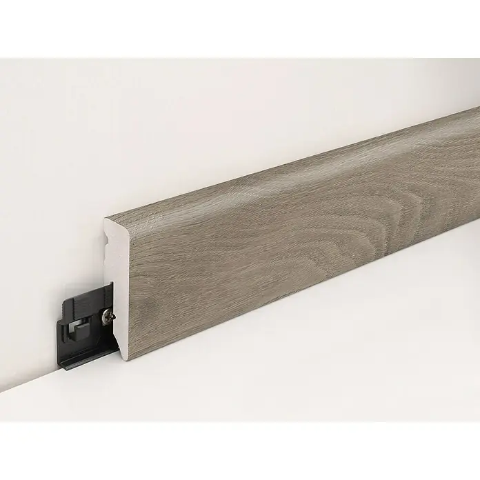 Profiles and more Sockelleiste KU51L Eiche Hartford - 240 cm x 15 mm x 50 mm - Eine Fußleiste in Holzoptik mit einem schwarzen Befestigungssystem, das an einer weißen Wand montiert ist, für ein elegantes Raumdesign.