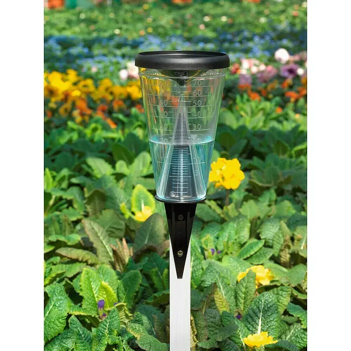 TFA Dostmann Regenmesser - Höhe: 305 mm - Nahaufnahme einer Regenmesser-Vorrichtung in einem Garten voller bunter Blumen, um die Niederschlagsmenge zu messen.