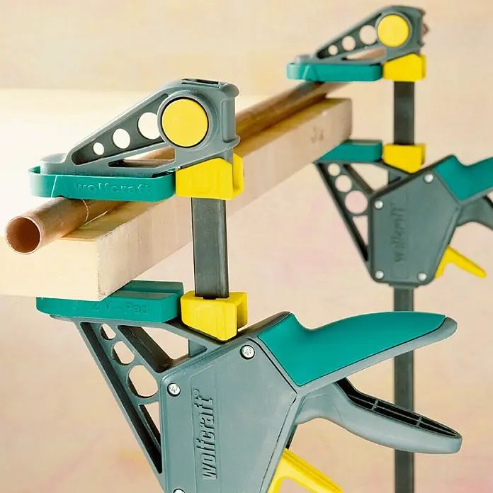 Wolfcraft Tornillo de apriete monomanual EHZ PRO - Anchura de sujeción: 915 mm, Fuerza de tensión: 1.200 N, Apertura máxima: 100 mm - Primer plano de dos abrazaderas de una mano Wolfcraft que sujetan un trozo de madera y una tubería de cobre. Ideal para proyectos de taller.