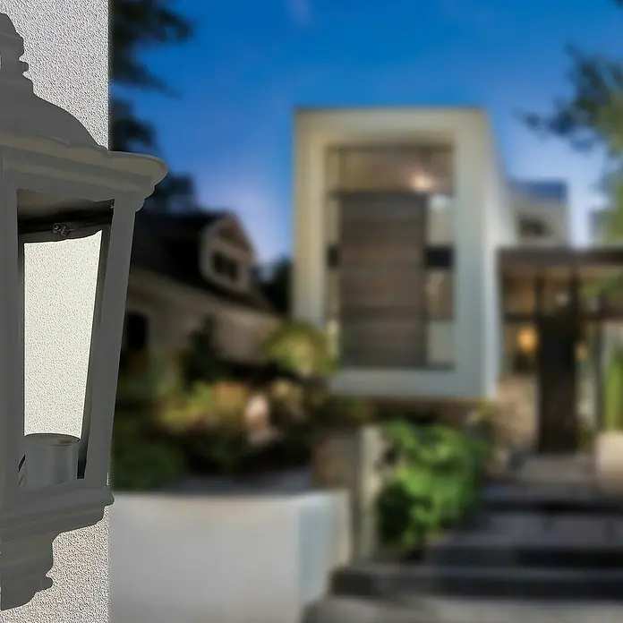 Panaled Vagg Aplique exterior - 60 W, 12,1 x 25,1 x 44 cm, Negro, IP44 - Primer plano de una lámpara de pared blanca frente a una casa. La luz es elegante y añade un toque de estilo al exterior.