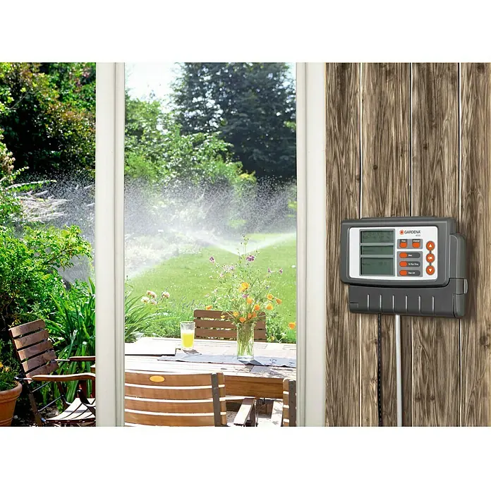 Gardena Sprinklersystem Programador 4030 Classic - Tiempo de riego: 1 min - 4 h - La imagen muestra un sistema de control de riego Gardena montado en una pared de madera, con vista a un jardín con aspersores a través de una ventana.