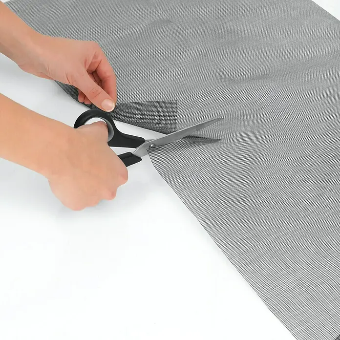Easy Maxx Insektenschutz-Netz - B x H: 130 x 150 cm, Montage Insektengitter: Magnetisch anheften - Nahaufnahme von Händen, die ein Stück graues Fliegengitter mit einer Schere auf einem weißen Tisch schneiden.