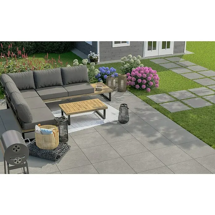 Terrassenfliese Garden Family Lightgrey - 60 x 60 x 2 cm, Hellgrau, Matt - Gartenmöbel-Set mit grauen Polstern und Holztischen auf einer grauen Terrasse, umgeben von Blumen und Grünpflanzen.