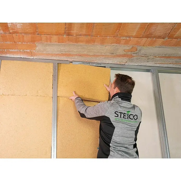 Steico Holzfaserdämmplatte flex 0381.200 x 625 mm, Stärke: 60 mm, Wärmeleitfähigkeit: 0,038 W/(mK), 7 Stk. - Ein Mann befestigt eine Steico Holzfaserplatte an einer Wand. Das Steico-Logo ist auf seiner Jacke sichtbar, was die Marke hervorhebt.