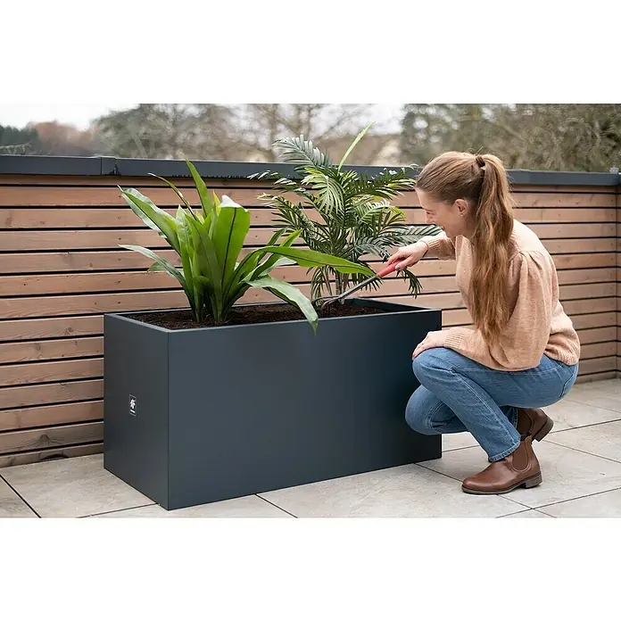 Herstera Garden Pflanzkasten Cube - Außenmaß (L x B x H): 100 x 50 x 50 cm, Metall, Anthrazit - Eine Frau arbeitet mit einer Harke in einem dunkelgrauen, rechteckigen Pflanzkasten mit grünen Pflanzen auf einer Terrasse.