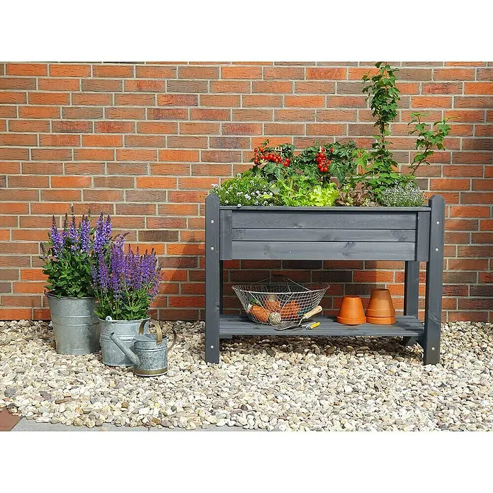 Promadino Kräuterhochbeet - 114 x 60 x 84,5 cm, Grau - Hochbeet aus Holz mit Pflanzen und Gartenzubehör vor einer Backsteinmauer.