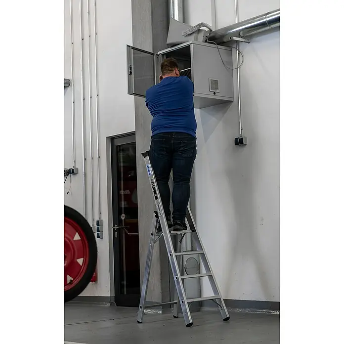 Krause Stabilo Stehleiter 4 Stufen - Anzahl Tritte: 4 Stk., Arbeitshöhe: 2,95 m, Belastbarkeit: 225 kg, Aluminium - Ein Mann steht auf einer silbernen Krause Leiter, um an einer grauen Wandmontagebox mit offenen Tür zu arbeiten. Silberne Rohre verlaufen über der Box.