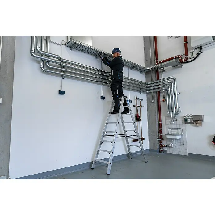 Krause Stabilo Doppel-Stehleiter 2 x 8 Stufen - Anzahl Tritte: 16 Stk., Arbeitshöhe: 3,4 m, Belastbarkeit: 225 kg, Aluminium - Ein Mann auf einer Leiter, der an Rohren in einem Gebäude arbeitet. Die Leiter ist silbern und der Mann trägt Arbeitskleidung.