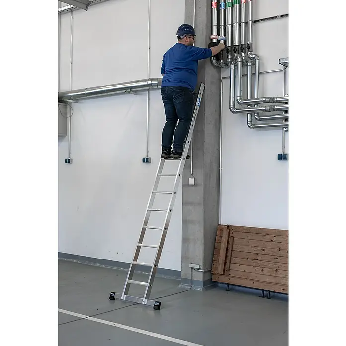 Krause Stabilo Anlegeleiter 12 Stufen - Anzahl Tritte: 12 Stk., Arbeitshöhe: 4,1 m, Belastbarkeit: 225 kg, Aluminium - Eine freistehende Aluminium-Leiter mit einer Person, die an einem Rohrsystem arbeitet, die die Vielseitigkeit und Sicherheit der Leiter hervorhebt.