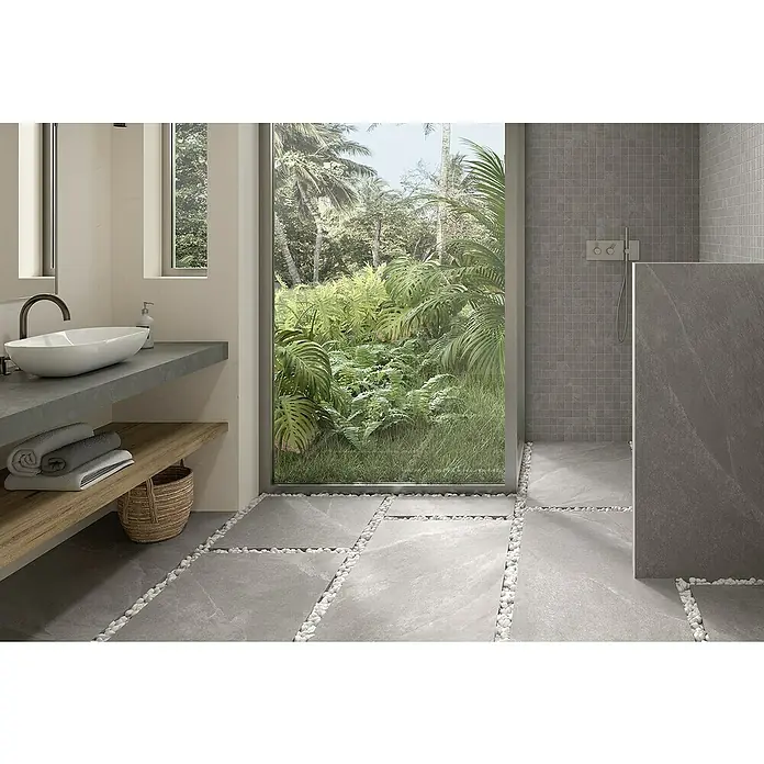 Momastela Feinsteinzeugfliese 2.0 Concept - 60 x 60 cm, Grigio - Helles Badezimmer mit Waschbecken, Holzbord und grauen Fliesen, die durch weiße Kieselsteine getrennt sind, sowie Blick auf üppiges Grün durch ein Fenster.