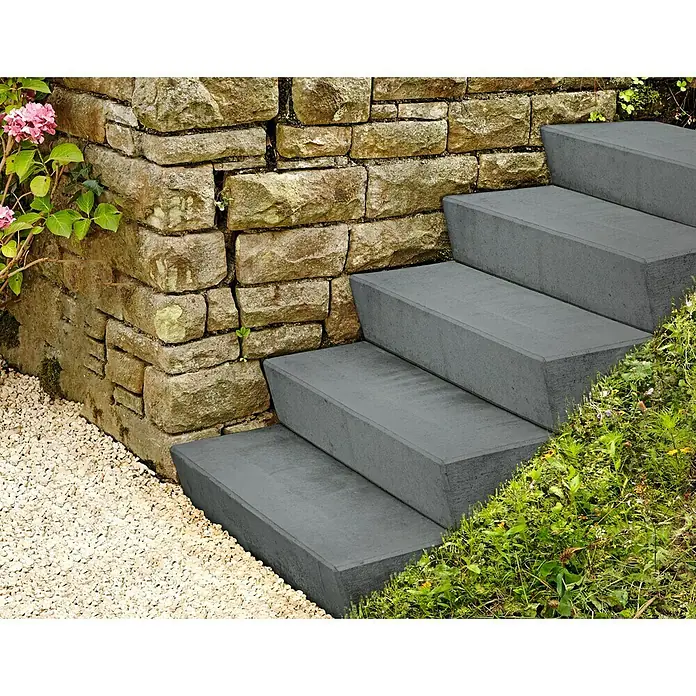 Christoph Blockstufe - Anthrazit, 50 x 32 x 16 cm, Beton - Betontreppen führen an einer steinernen Stützmauer entlang, umgeben von Kies und üppigem Grün. Die Treppe ist modern und schlicht gestaltet.