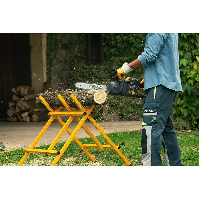 Stiga Akku-Kettensäge CS 700E (14") - 48 V, Ohne Akku, Schwertlänge: 35 cm - Ein Mann sägt Holz mit einer STIGA Kettensäge auf einem gelben Sägebock, umgeben von herumfliegenden Holzspänen.