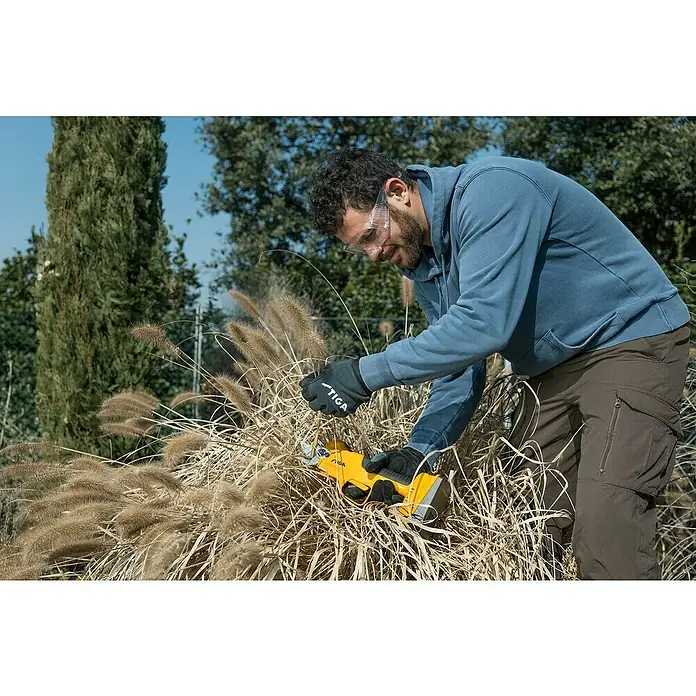 Stiga Akku-Astschere SC 100E - 20 V, 1 Akku, Max. Aststärke: 30 mm - Ein Mann, der mit der gelben STIGA Akku-Heckenschere Gräser schneidet, trägt dabei Schutzbrille und Handschuhe für sicheres Arbeiten im Garten.