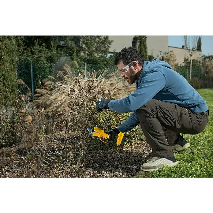 Stiga Akku-Astschere SC 100E - 20 V, 1 Akku, Max. Aststärke: 30 mm - Ein Mann kniet im Garten und schneidet mit einer STIGA Gartenschere eine Pflanze. Er trägt eine Schutzbrille und Handschuhe.
