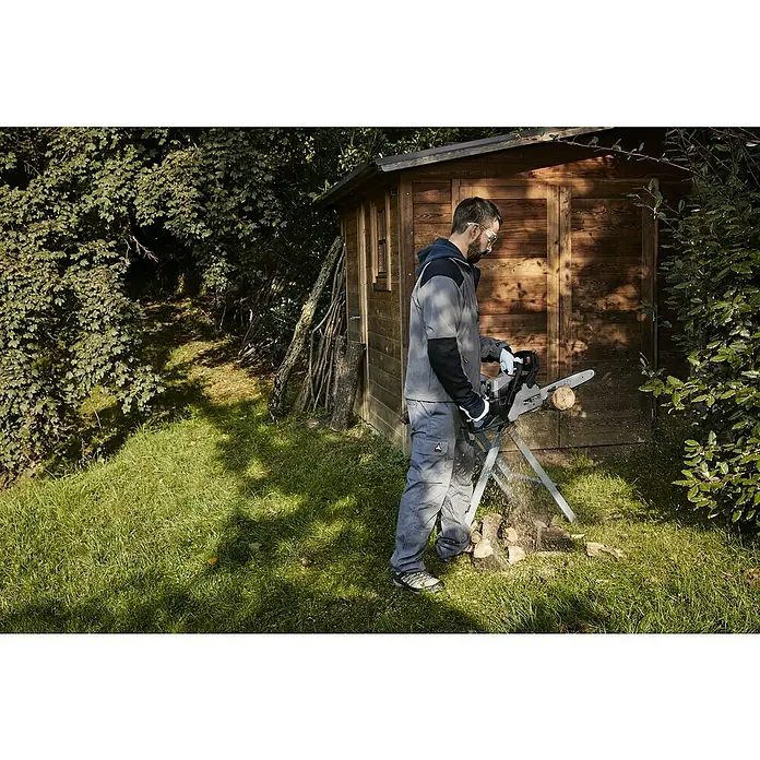 Alpina Benzin-Kettensäge ACS 39 - 1,2 kW, 37,2 cm³, Schwertlänge: 35 cm - Ein Mann benutzt eine Kettensäge, um Holz zu schneiden, während er Schutzbrille und Handschuhe trägt. Im Hintergrund befindet sich ein Holzschuppen.