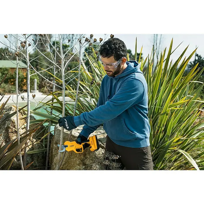 Stiga Akku-Astschere SC 100E - 20 V, 1 Akku, Max. Aststärke: 30 mm - Ein Mann benutzt eine gelbe STIGA Astschere im Garten, um Äste zu schneiden, während er Schutzbrille und Handschuhe trägt.