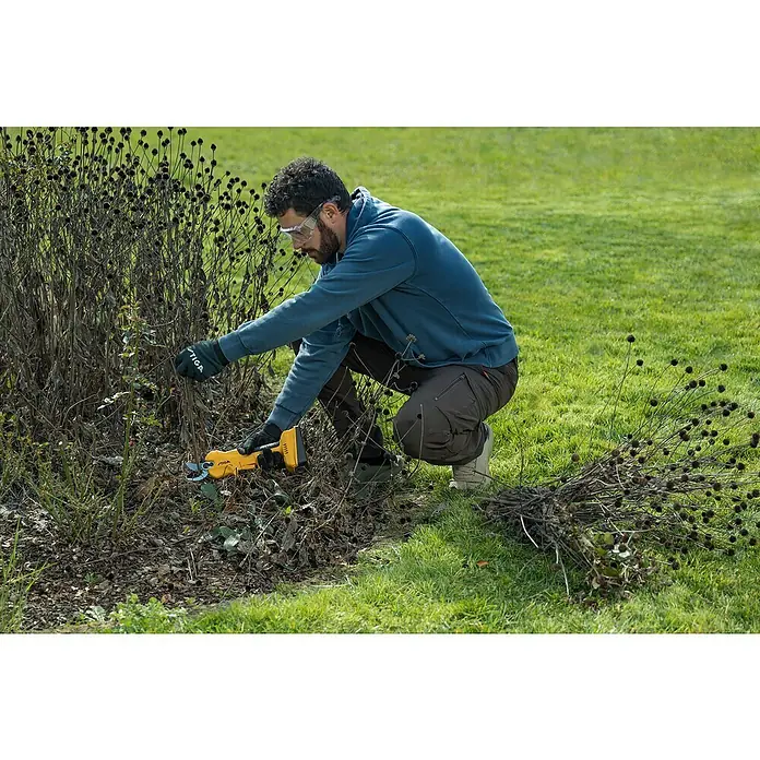 Stiga Akku-Astschere SC 100E - 20 V, 1 Akku, Max. Aststärke: 30 mm - Ein Mann mit Schutzbrille und Handschuhen schneidet mit einer gelben STIGA Astschere eine Pflanze im Garten.