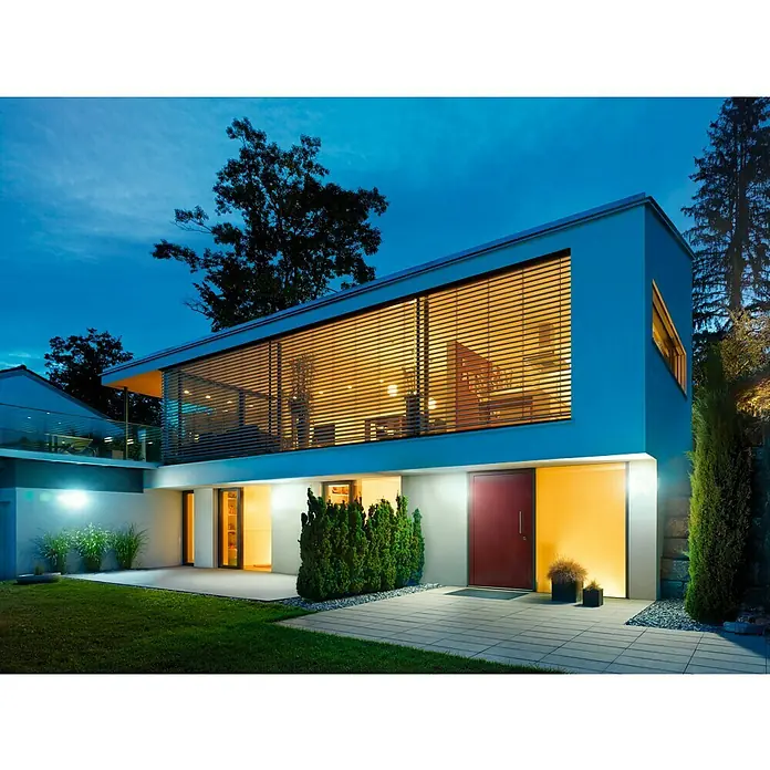 Steinel LED-Sensor-Strahler XLED home Curved S anthrazit - 9,3 W, L x B x H: 16 x 10,7 x 12 cm, Anthrazit, IP44 - Modernes Haus mit roter Tür und großen Fenstern mit Jalousien in der Abenddämmerung. Die Beleuchtung betont die architektonischen Details.
