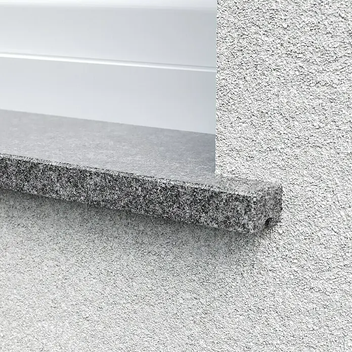 Fensterbank Impakt softgebürstet - 113 x 30 x 2 cm, Anthrazit, Naturstein - Nahaufnahme einer Fensterbank aus Granit, die in eine strukturierte, weiße Wand eingelassen ist, mit einem Rollladen im Hintergrund.