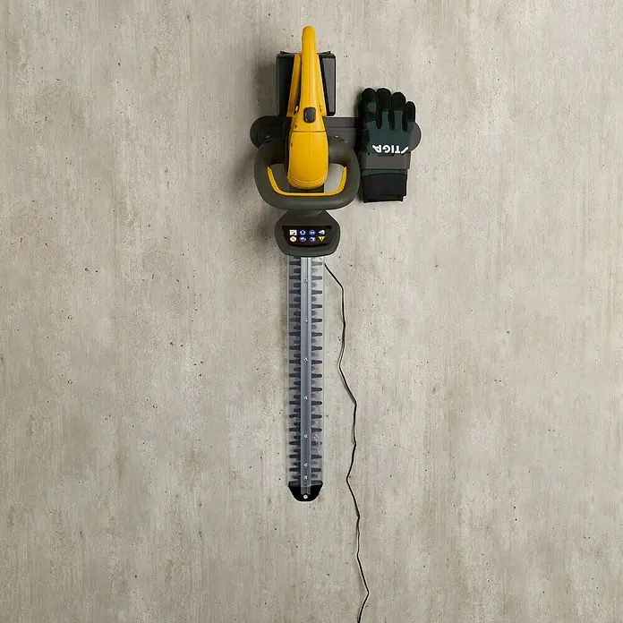 Stiga Akku-Heckenschere HT 300E - 2 Akkus, Schnittlänge: 54 cm - Eine gelbe Heckenschere mit Akku hängt mit einem Arbeitshandschuh an einer Halterung an der Wand.