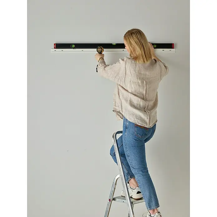 Elfa Classic by Elfa Aufhängeschiene - Länge: 936 mm, Metall, Weiß - Eine Frau auf einer Leiter montiert mit einem Akkuschrauber eine lange, schwarze Wasserwaage an einer weißen Wand.