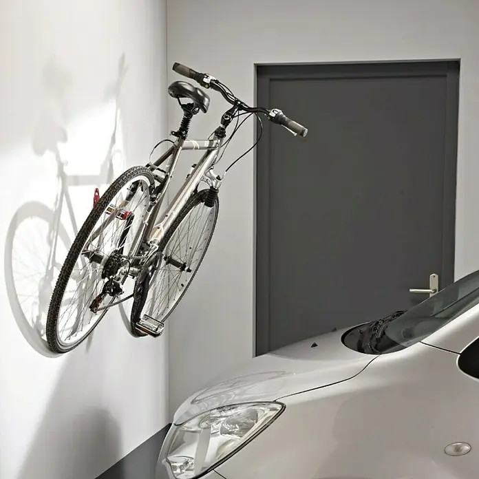 Mottez Gancho de pared para bicicletas B215V - Acero - Una bicicleta plateada montada en una pared blanca en un garaje, al lado de un coche. La bici está almacenada de manera segura.