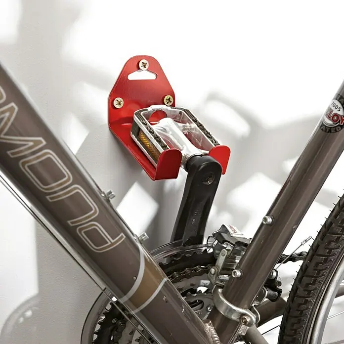 Mottez Gancho de pared para bicicletas B215V - Acero - La bicicleta está fijada a la pared con un soporte de pared rojo. Los pedales de la bicicleta se mantienen en su lugar por el soporte.